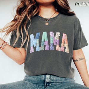 May include: Dark grey t-shirt with the word "MAMA" in patchwork letters. The letters are made of various patterned fabrics, including gingham, floral, and polka dots. The word "PEPPER" is in the top right corner.
