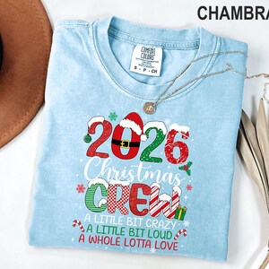 以下が含まれることがあります： 2026 Christmas CREW A LITTLE BIT CRAZY A LITTLE BIT LOUD A WHOLE LOTTA LOVEと書かれたグラフィックプリント入りのライトブルーの半袖Tシャツ。赤、緑、白のクリスマス柄、サンタの帽子、キャンディケインのデザインが施されています。