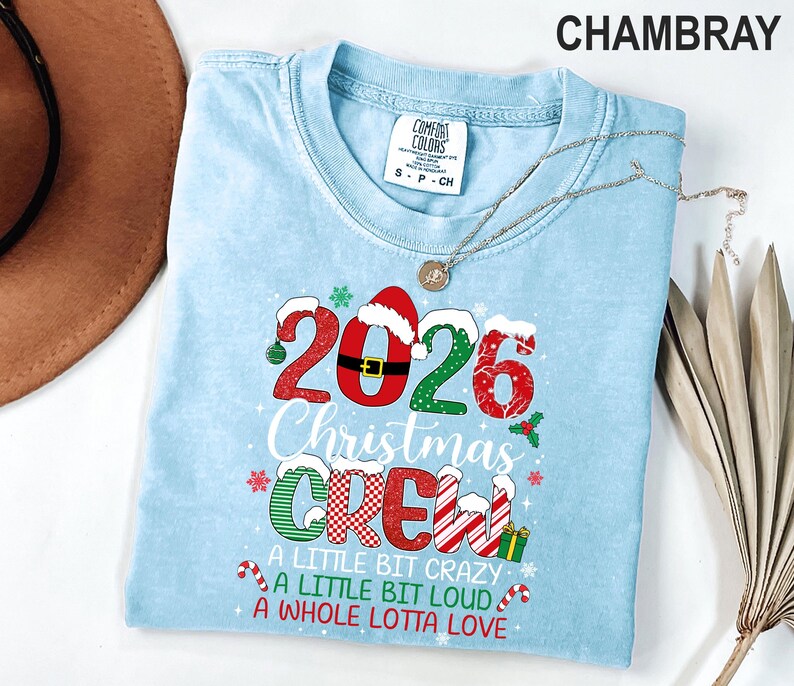 以下が含まれることがあります： 2026 Christmas CREW A LITTLE BIT CRAZY A LITTLE BIT LOUD A WHOLE LOTTA LOVEと書かれたグラフィックプリント入りのライトブルーの半袖Tシャツ。赤、緑、白のクリスマス柄、サンタの帽子、キャンディケインのデザインが施されています。