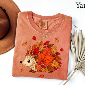 May include: A yam-colored crew neck t-shirt featuring a graphic of a hedgehog whose back is covered in various autumn-colored leaves including red, orange, and brown. The shirt has a Comfort Colors brand label at the inner neckline with size S.