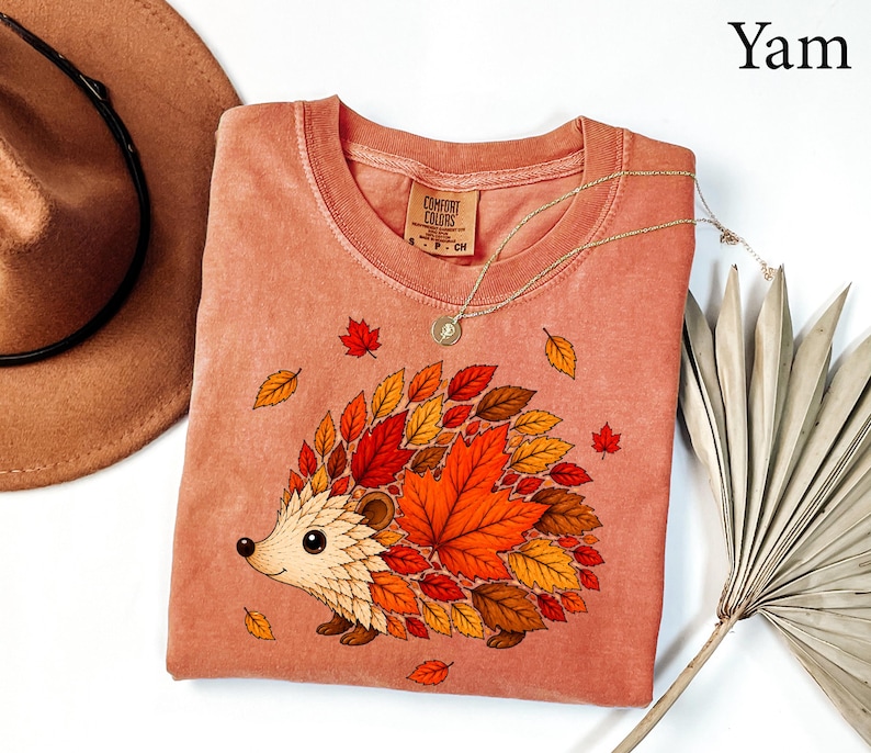 May include: A yam-colored crew neck t-shirt featuring a graphic of a hedgehog whose back is covered in various autumn-colored leaves including red, orange, and brown. The shirt has a Comfort Colors brand label at the inner neckline with size S.
