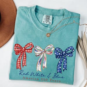 May include: A teal short-sleeved t-shirt featuring three bows in red gingham, white with gold accents, and blue with white stars. Below the bows, the text reads Red White & Blue America 250 Years. The shirt has a crew neckline and a Comfort Colors label.