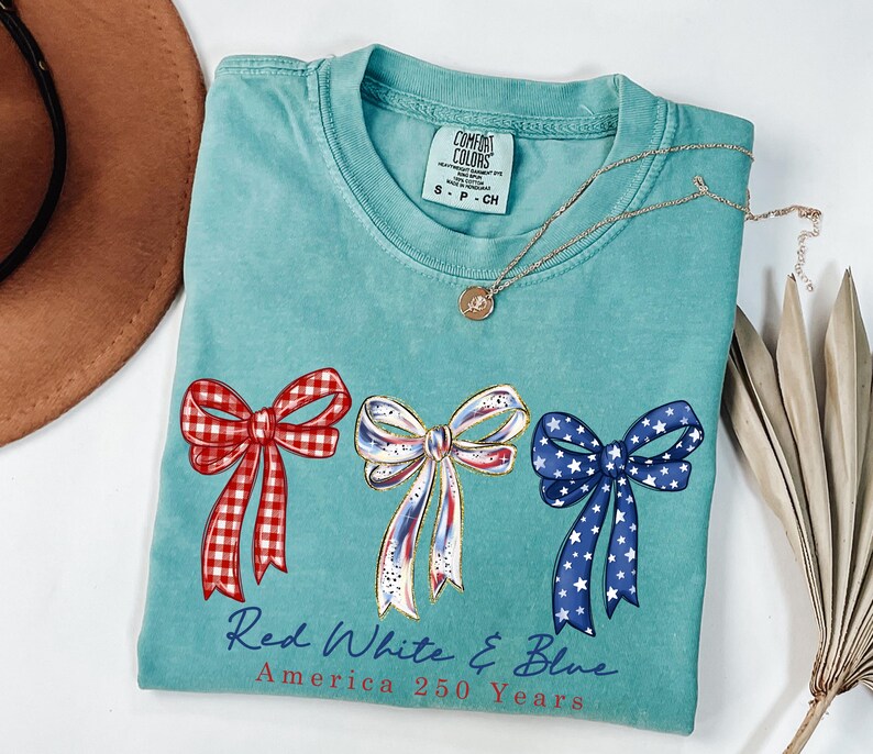 May include: A teal short-sleeved t-shirt featuring three bows in red gingham, white with gold accents, and blue with white stars. Below the bows, the text reads Red White & Blue America 250 Years. The shirt has a crew neckline and a Comfort Colors label.