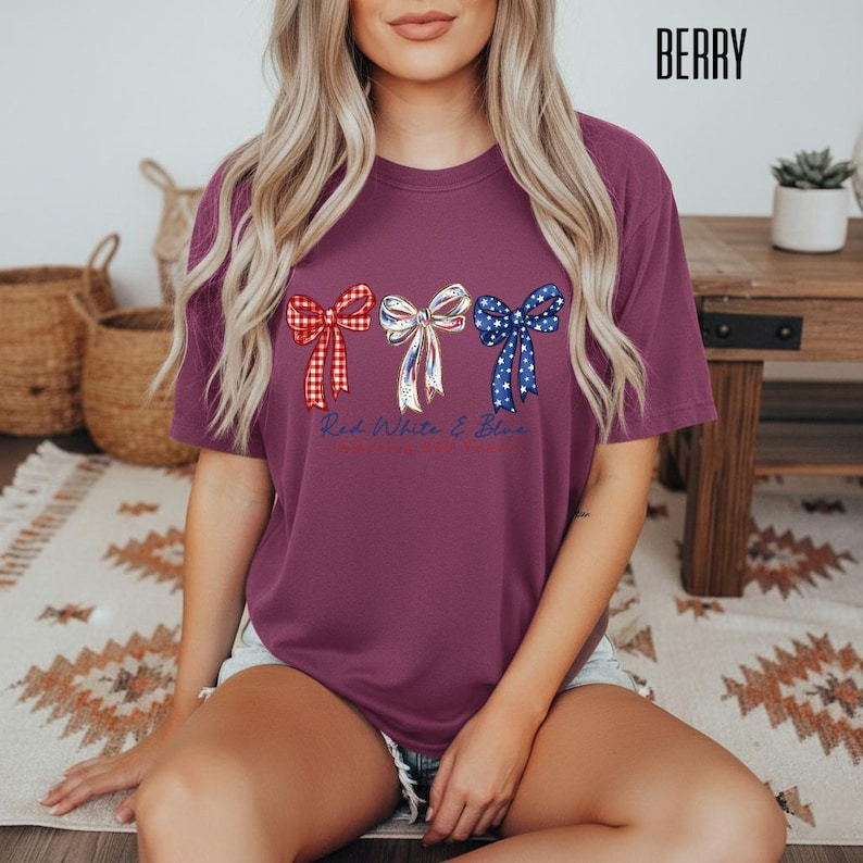 May include: A berry-colored short-sleeve t-shirt featuring three decorative bows printed across the chest. The left bow is red and white checkered, the center bow is white with blue and red accents, and the right bow is blue with white stars.