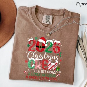 以下が含まれることがあります： エスプレッソカラーのクルーネックTシャツ。2026 Christmas Crew, A Little Bit Crazy, A Little Bit Loud, A Whole Lotta Loveという文字がプリントされています。デザインには、クリスマスの模様、サンタの帽子、キャンディケインがあしらわれています。