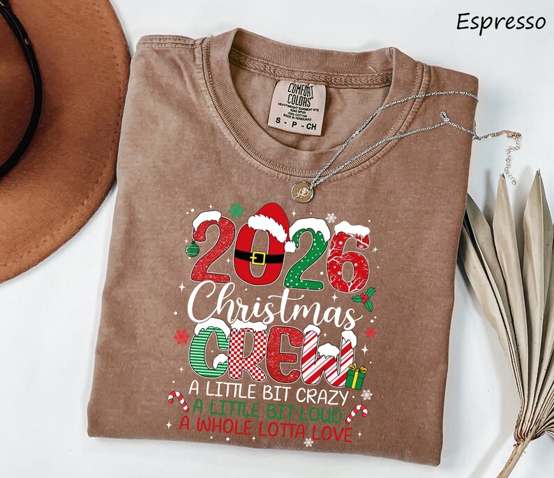 以下が含まれることがあります： エスプレッソカラーのクルーネックTシャツ。2026 Christmas Crew, A Little Bit Crazy, A Little Bit Loud, A Whole Lotta Loveという文字がプリントされています。デザインには、クリスマスの模様、サンタの帽子、キャンディケインがあしらわれています。