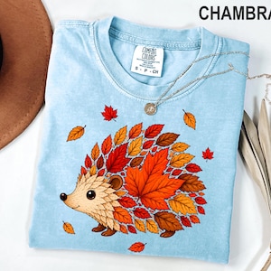 May include: A light blue t-shirt with a graphic of a hedgehog whose back is covered in red, orange, and yellow autumn leaves. The shirt features a crew neckline and a white interior label with the text Comfort Colors, S P CH, and 100% cotton.