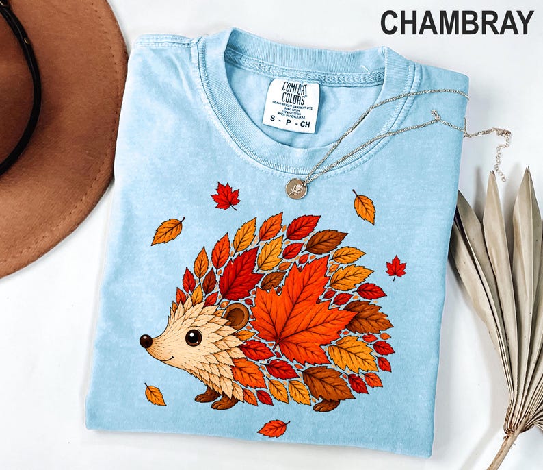 May include: A light blue t-shirt with a graphic of a hedgehog whose back is covered in red, orange, and yellow autumn leaves. The shirt features a crew neckline and a white interior label with the text Comfort Colors, S P CH, and 100% cotton.