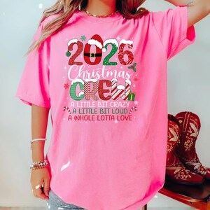 以下が含まれることがあります： 2026 Christmas CREW, A LITTLE BIT CRAZY, A LITTLE BIT LOUD, A WHOLE LOTTA LOVEという文字と、サンタの帽子やキャンディケイン、ヒイラギなどのクリスマスモチーフがプリントされた、鮮やかなピンク色の半袖Tシャツ。