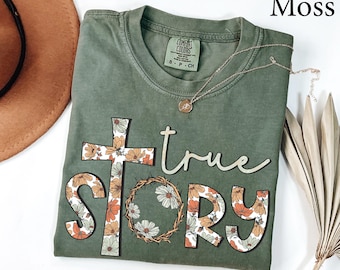 Puede incluir: Camiseta verde musgo con una cruz floral y las palabras "true story" en una fuente retro. Las letras y la cruz están decoradas con flores naranjas, blancas y marrones. Un collar dorado con un colgante está drapeado sobre la camiseta.