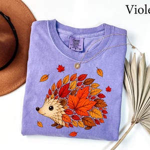 May include: A violet short-sleeved t-shirt with a graphic print of a hedgehog whose back is covered in colorful autumn leaves in shades of red, orange, and yellow. The shirt features a crew neckline and a Comfort Colors brand label at the neck.