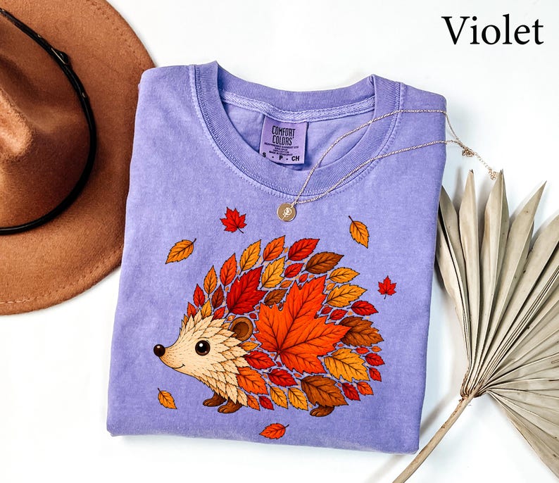 May include: A violet short-sleeved t-shirt with a graphic print of a hedgehog whose back is covered in colorful autumn leaves in shades of red, orange, and yellow. The shirt features a crew neckline and a Comfort Colors brand label at the neck.