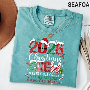以下が含まれることがあります： シーフォームグリーンのTシャツ。2026 CHRISTMAS CREW A LITTLE BIT CRAZY A LITTLE BIT LOUD A WHOLE LOTTA LOVEという文字がプリントされています。数字と文字は赤、緑、白の模様、サンタの帽子、雪のモチーフで装飾されています。