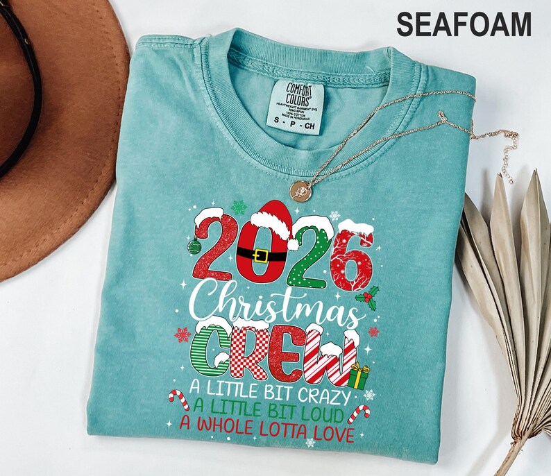 以下が含まれることがあります： シーフォームグリーンのTシャツ。2026 CHRISTMAS CREW A LITTLE BIT CRAZY A LITTLE BIT LOUD A WHOLE LOTTA LOVEという文字がプリントされています。数字と文字は赤、緑、白の模様、サンタの帽子、雪のモチーフで装飾されています。
