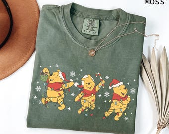 Puede incluir: Una camiseta verde musgo con un gráfico de tres personajes de Winnie the Pooh adornados con luces navideñas, gorros de Papá Noel y bufandas. La camiseta tiene cuello redondo y mangas cortas. Un collar dorado está drapeado sobre la camiseta.