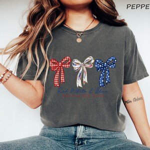 May include: A charcoal gray short-sleeve t-shirt featuring a graphic of three bows in red gingham, white with blue and red accents, and blue with white stars. The text "Red White & Blue" is printed in blue cursive script below the bows.