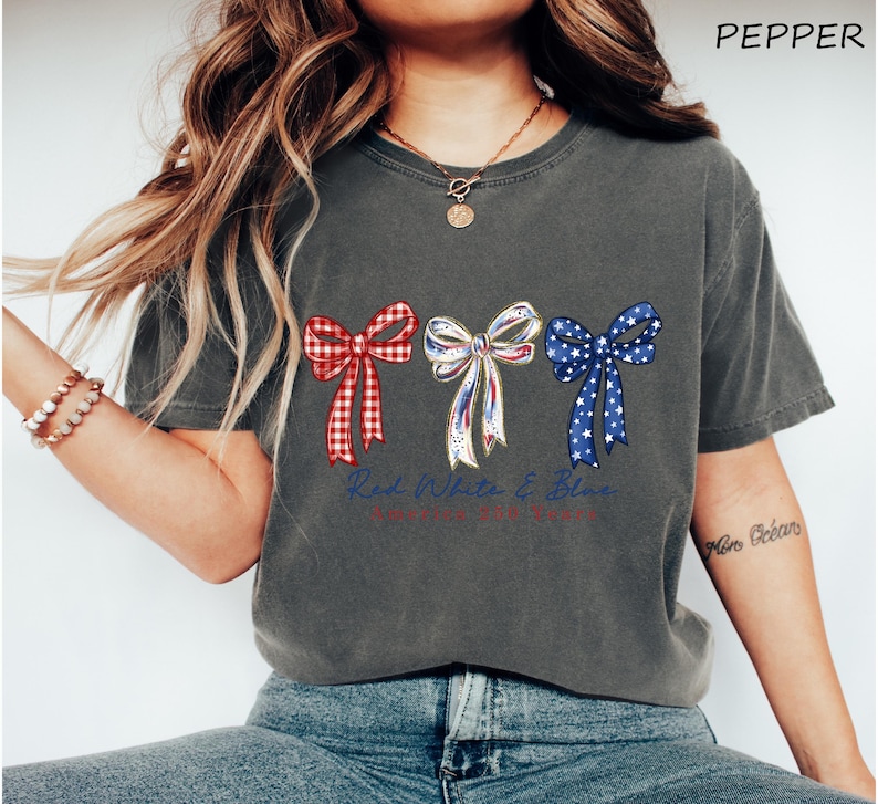 May include: A charcoal gray short-sleeve t-shirt featuring a graphic of three bows in red gingham, white with blue and red accents, and blue with white stars. The text "Red White & Blue" is printed in blue cursive script below the bows.