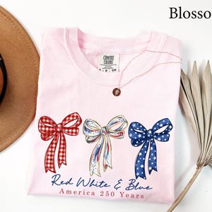 May include: A light pink t-shirt featuring three illustrated bows in red gingham, white with blue and red accents, and blue with white stars. Below the bows, the text reads Red White & Blue and America 250 Years in blue and red script lettering.