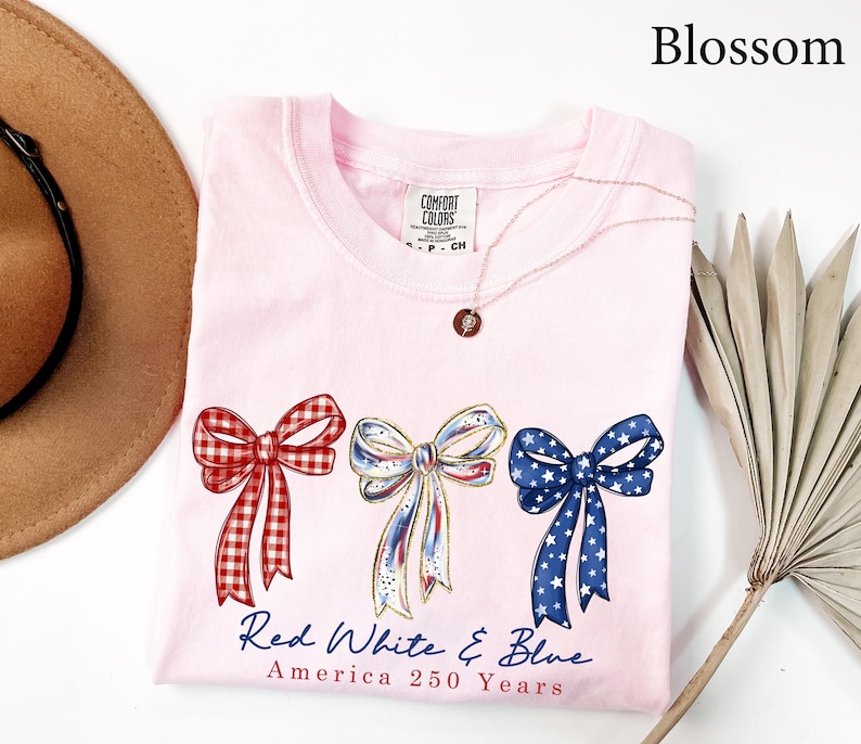 May include: A light pink t-shirt featuring three illustrated bows in red gingham, white with blue and red accents, and blue with white stars. Below the bows, the text reads Red White & Blue and America 250 Years in blue and red script lettering.