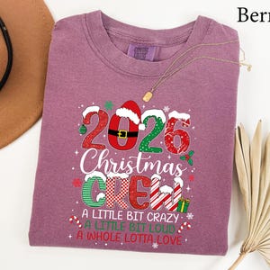 以下が含まれることがあります： 2026 Christmas CREW, A LITTLE BIT CRAZY, A LITTLE BIT LOUD, A WHOLE LOTTA LOVEというテキストがプリントされたベリー色のTシャツ。赤、緑、白のパターン、サンタの帽子、小さなクリスマスのアイコンがデザインされています。