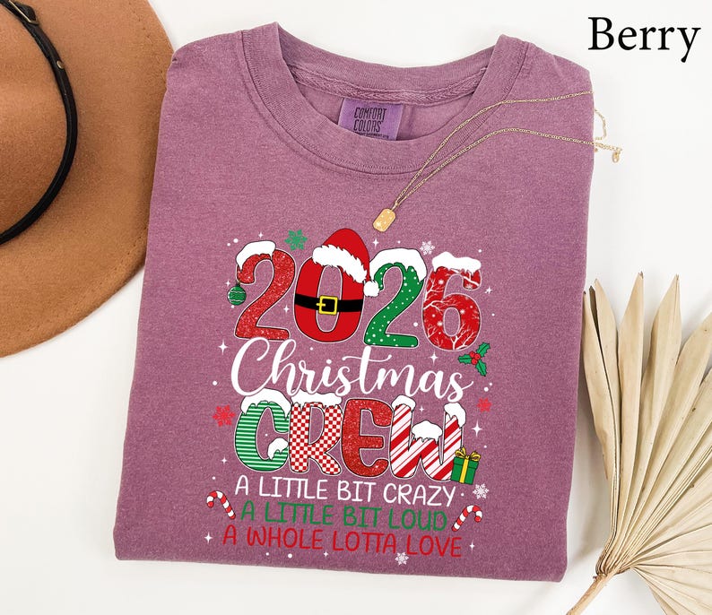 以下が含まれることがあります： 2026 Christmas CREW, A LITTLE BIT CRAZY, A LITTLE BIT LOUD, A WHOLE LOTTA LOVEというテキストがプリントされたベリー色のTシャツ。赤、緑、白のパターン、サンタの帽子、小さなクリスマスのアイコンがデザインされています。