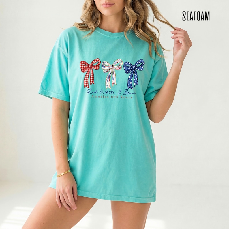 May include: A seafoam green short-sleeve t-shirt featuring a graphic of three bows in red gingham, white with blue stars, and blue with white stars. Below the bows, the text reads Red White & Blue America 250 Years in a cursive font.