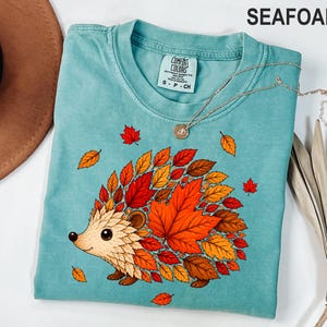 May include: A seafoam green t-shirt featuring a graphic of a hedgehog whose back is covered in vibrant red, orange, and yellow autumn leaves. The shirt has a crew neckline and a Comfort Colors brand label on the inside collar with size S-CH.