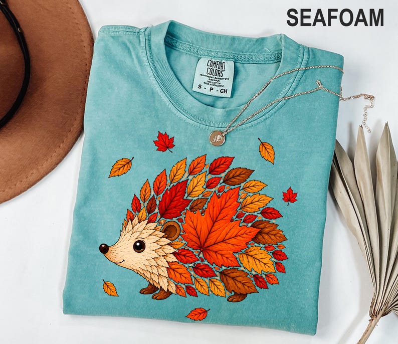 May include: A seafoam green t-shirt featuring a graphic of a hedgehog whose back is covered in vibrant red, orange, and yellow autumn leaves. The shirt has a crew neckline and a Comfort Colors brand label on the inside collar with size S-CH.