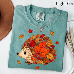May include: A light green t-shirt featuring a graphic of a hedgehog whose body is composed of various autumn-colored leaves in shades of red, orange, and brown. The shirt has a crew neckline and a Comfort Colors brand label on the inside collar.