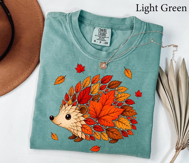 May include: A light green t-shirt featuring a graphic of a hedgehog whose body is composed of various autumn-colored leaves in shades of red, orange, and brown. The shirt has a crew neckline and a Comfort Colors brand label on the inside collar.