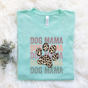 May include: A light blue t-shirt with the text "DOG MAMA" repeated in a stacked design with a leopard print paw print in the center. The text is in a vintage style font.