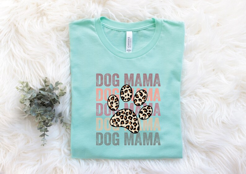 May include: A light blue t-shirt with the text "DOG MAMA" repeated in a stacked design with a leopard print paw print in the center. The text is in a vintage style font.