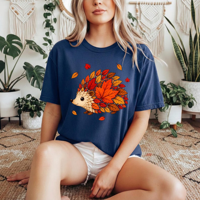 May include: A navy blue t-shirt featuring a graphic illustration of a hedgehog whose back is covered in vibrant orange, red, and yellow autumn leaves. The hedgehog has a light tan face and a small black nose, with scattered leaves around it.