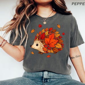 May include: A charcoal gray t-shirt featuring a graphic of a hedgehog whose body is composed of vibrant orange, red, and yellow autumn leaves. The word PEPPER is printed in the upper right corner of the image in a simple black font.