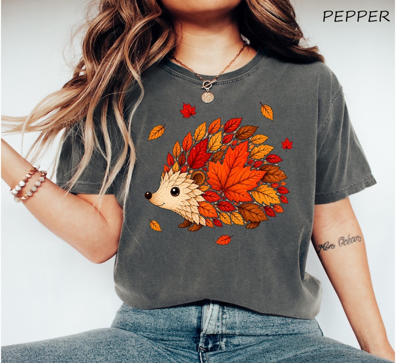 May include: A charcoal gray t-shirt featuring a graphic of a hedgehog whose body is composed of vibrant orange, red, and yellow autumn leaves. The word PEPPER is printed in the upper right corner of the image in a simple black font.