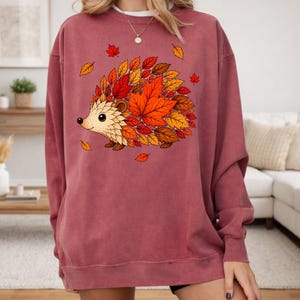 May include: A dusty rose crewneck sweatshirt featuring a central graphic of a hedgehog whose body is composed of vibrant autumn leaves in shades of red, orange, and brown. The design includes scattered falling leaves around the hedgehog.