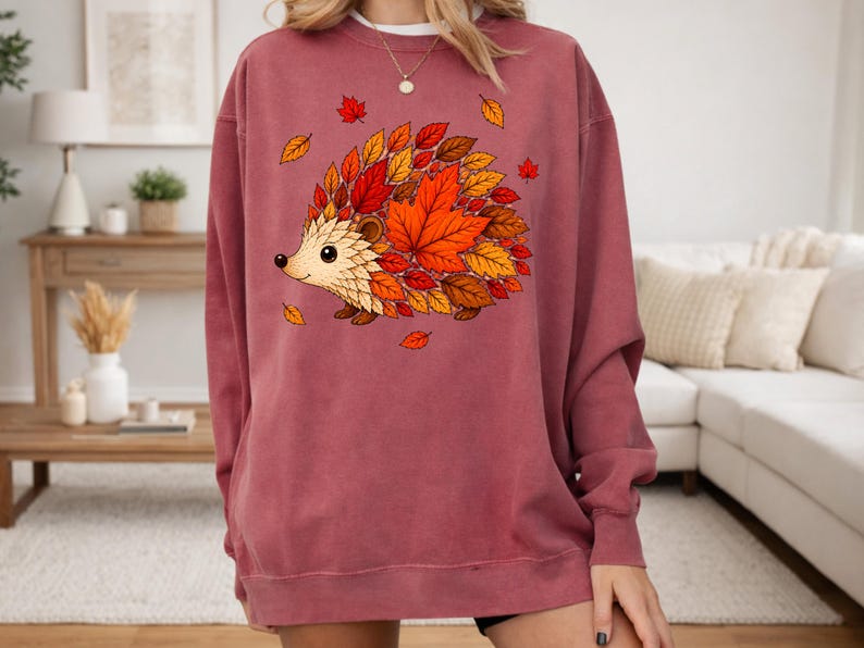 May include: A dusty rose crewneck sweatshirt featuring a central graphic of a hedgehog whose body is composed of vibrant autumn leaves in shades of red, orange, and brown. The design includes scattered falling leaves around the hedgehog.