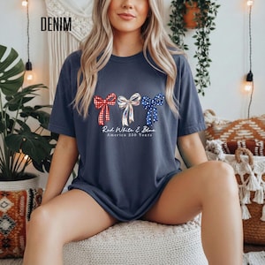 May include: A dark blue t-shirt featuring three decorative bows in red, white, and blue patterns. Below the bows, the text reads Red White & Blue America 250 Years. The shirt has a crew neckline and short sleeves, presented in a casual, relaxed fit.