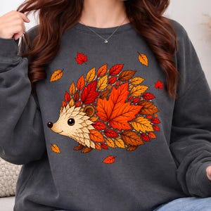 May include: A charcoal gray crewneck sweatshirt featuring a graphic illustration of a hedgehog. The hedgehog's body is composed of various autumn-colored leaves in shades of red, orange, and yellow. The design is centered on the front of the garment.