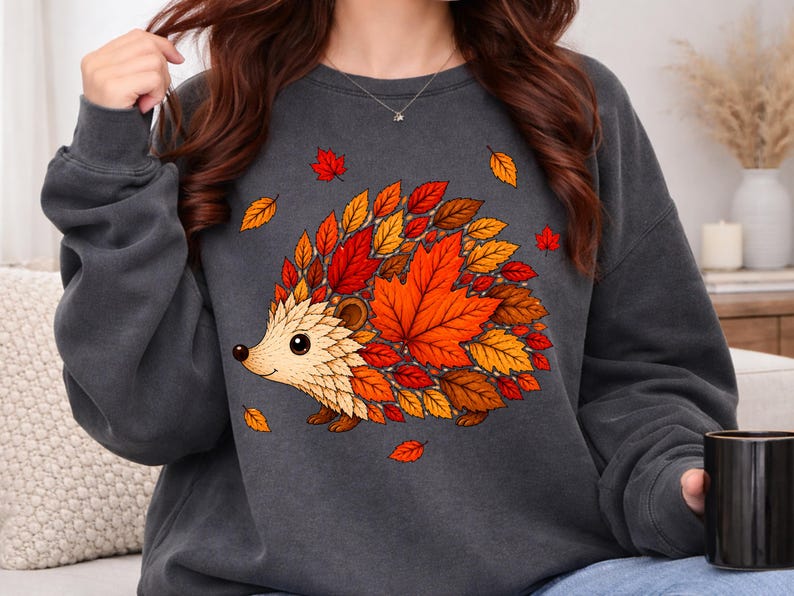 May include: A charcoal gray crewneck sweatshirt featuring a graphic illustration of a hedgehog. The hedgehog's body is composed of various autumn-colored leaves in shades of red, orange, and yellow. The design is centered on the front of the garment.