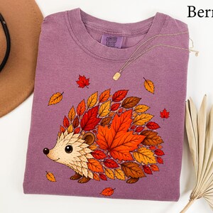 May include: A berry-colored t-shirt featuring a graphic of a hedgehog whose back is composed of various autumn leaves in shades of orange, red, and brown. The shirt has a crew neckline and a Comfort Colors brand label printed on the inner collar.