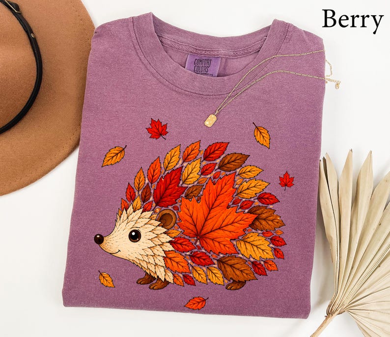 May include: A berry-colored t-shirt featuring a graphic of a hedgehog whose back is composed of various autumn leaves in shades of orange, red, and brown. The shirt has a crew neckline and a Comfort Colors brand label printed on the inner collar.