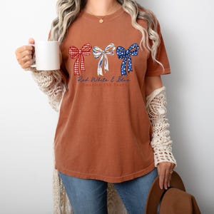 May include: A rust-colored short-sleeve t-shirt featuring three decorative bows in red gingham, white with blue accents, and blue with white stars. Below the bows, the text reads Red White & Blue America 250 Years in a cursive blue font.
