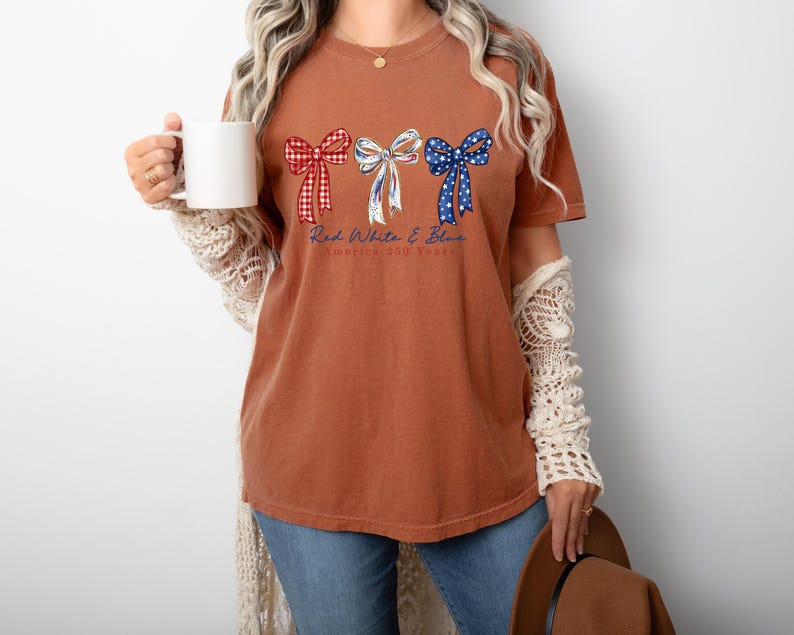 May include: A rust-colored short-sleeve t-shirt featuring three decorative bows in red gingham, white with blue accents, and blue with white stars. Below the bows, the text reads Red White & Blue America 250 Years in a cursive blue font.