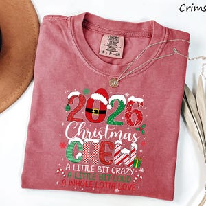 以下が含まれることがあります： 2026 Christmas Crew, A Little Bit Crazy, A Little Bit Loud, A Whole Lotta Loveというテキストがプリントされたクリムゾンカラーの半袖Tシャツ。デザインには、サンタの帽子、キャンディケイン、赤、白、緑のホリデーパターンが含まれています。