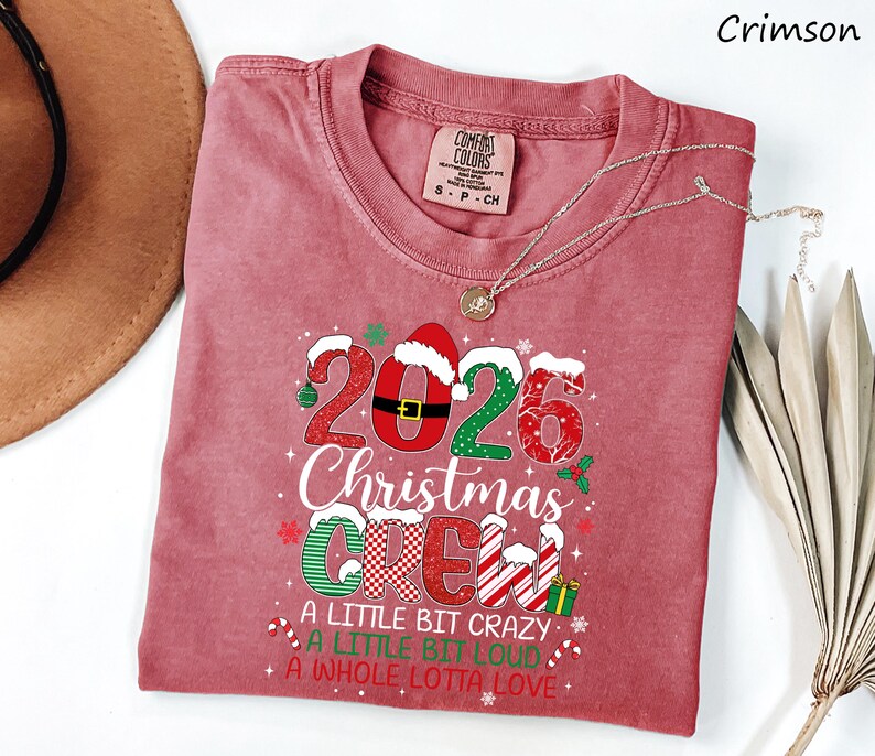 以下が含まれることがあります： 2026 Christmas Crew, A Little Bit Crazy, A Little Bit Loud, A Whole Lotta Loveというテキストがプリントされたクリムゾンカラーの半袖Tシャツ。デザインには、サンタの帽子、キャンディケイン、赤、白、緑のホリデーパターンが含まれています。