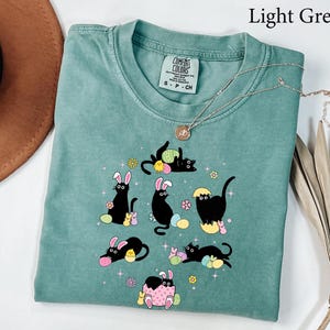 May include: Light green t-shirt with a crew neck and a graphic of black cats with bunny ears and Easter eggs. The text "Light Green" is in the upper right corner. A gold necklace is draped across the shirt.