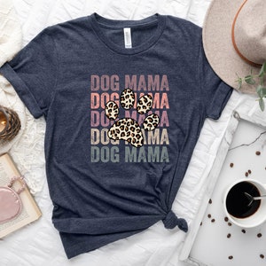 May include: A navy blue t-shirt with the text "DOG MAMA" repeated five times in a vintage style font. A leopard print paw print is centered on the shirt.