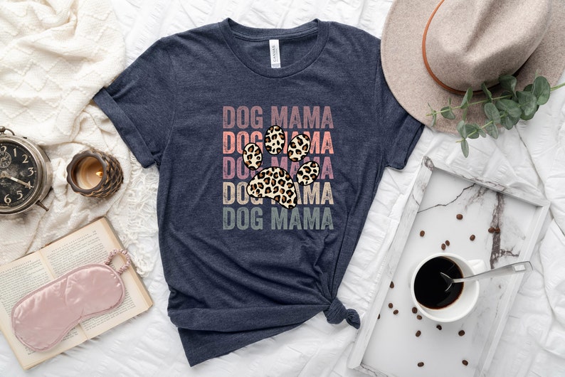 May include: A navy blue t-shirt with the text "DOG MAMA" repeated five times in a vintage style font. A leopard print paw print is centered on the shirt.