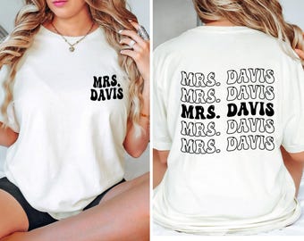 May include: A cream-colored short-sleeve t-shirt featuring the black text "MRS. DAVIS" on the left chest. The back of the shirt displays the text "MRS. DAVIS" repeated five times in a stacked, wavy black font with a solid fill.